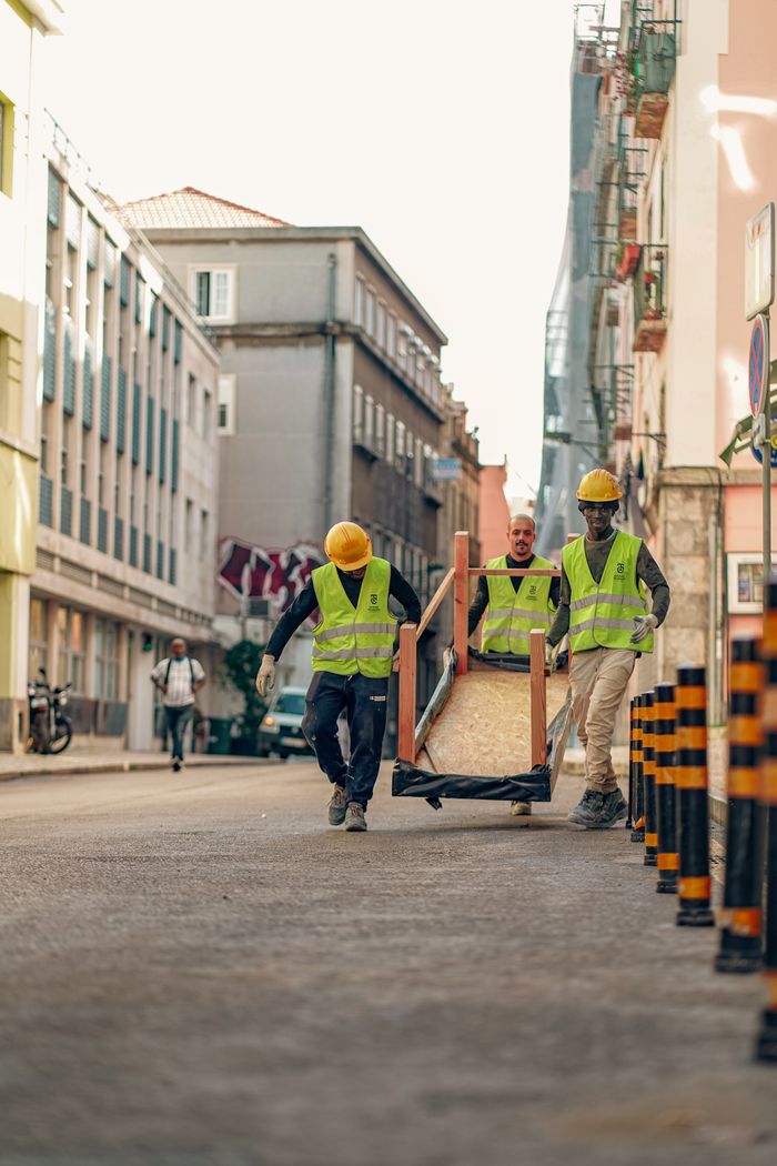 Three construction workers in safety vests transport heavy equipment on an urban street.