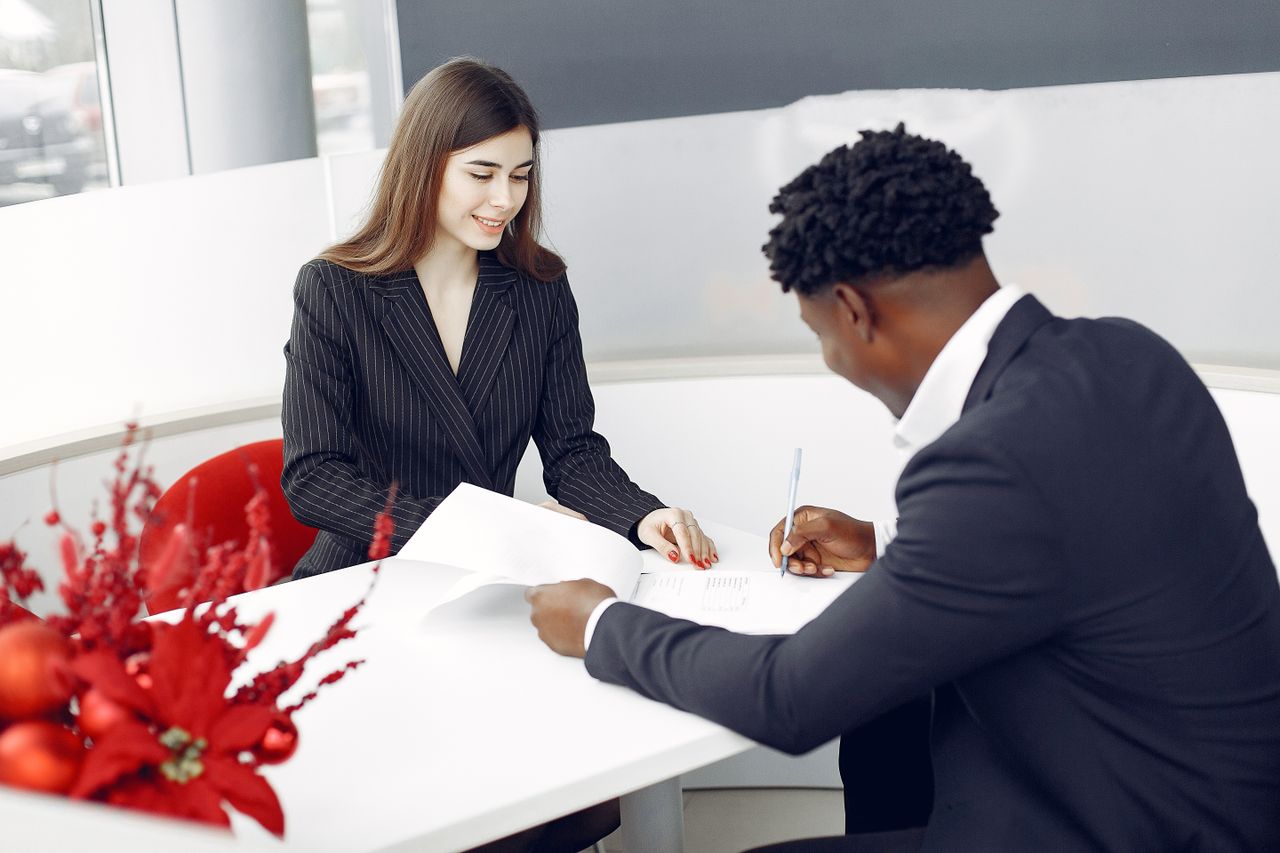 Positive stylish multiethnic buyer in formal classy suit and agent signing papers in modern light creative office in daylight and smiling happily